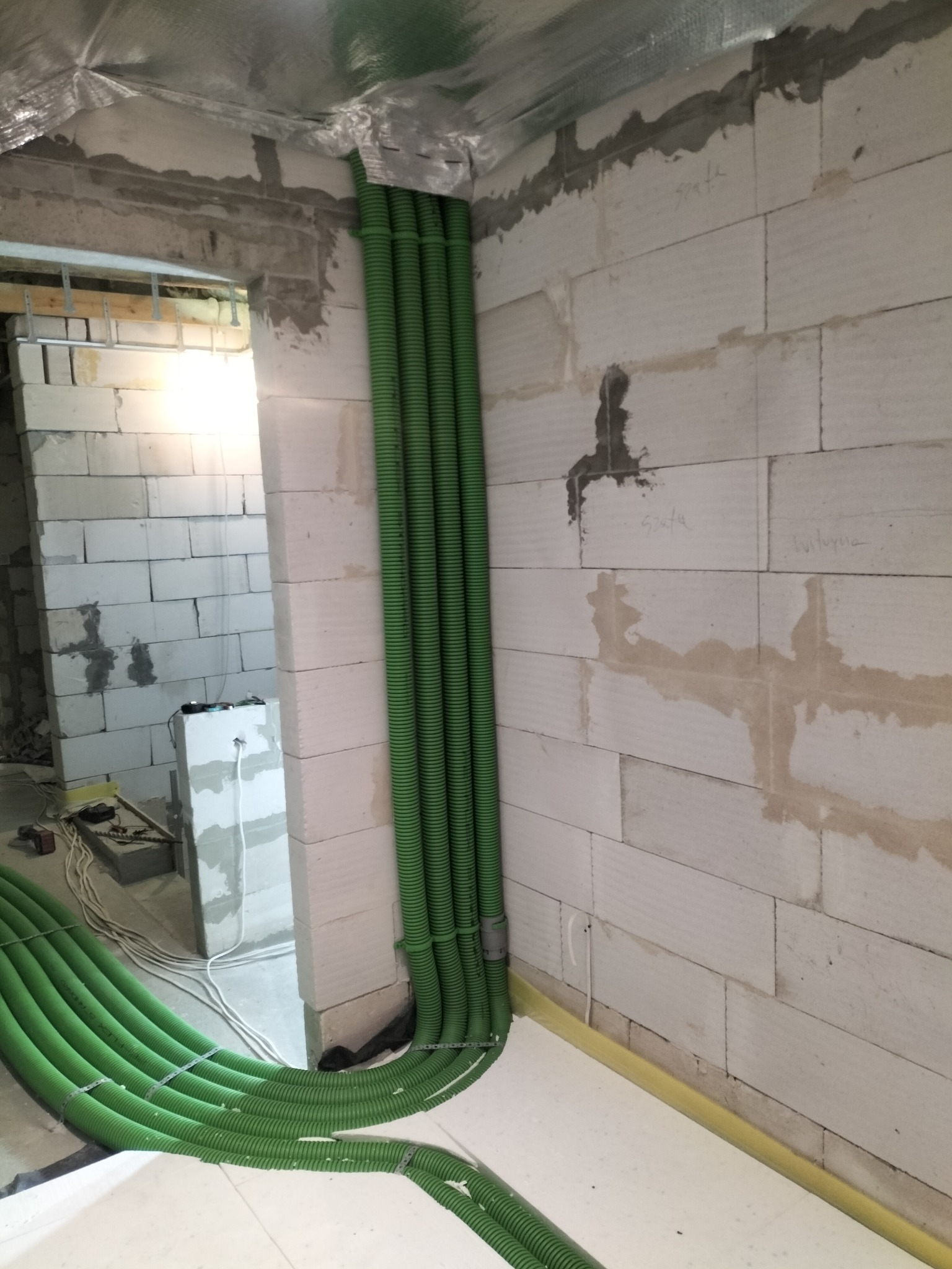 Zielone rury instalacyjne, prowadzone wzdłuż surowej ściany z betonu komórkowego, zabezpieczone srebrną folią. Widoczne mocowania i fragment podłogi z izolacją.