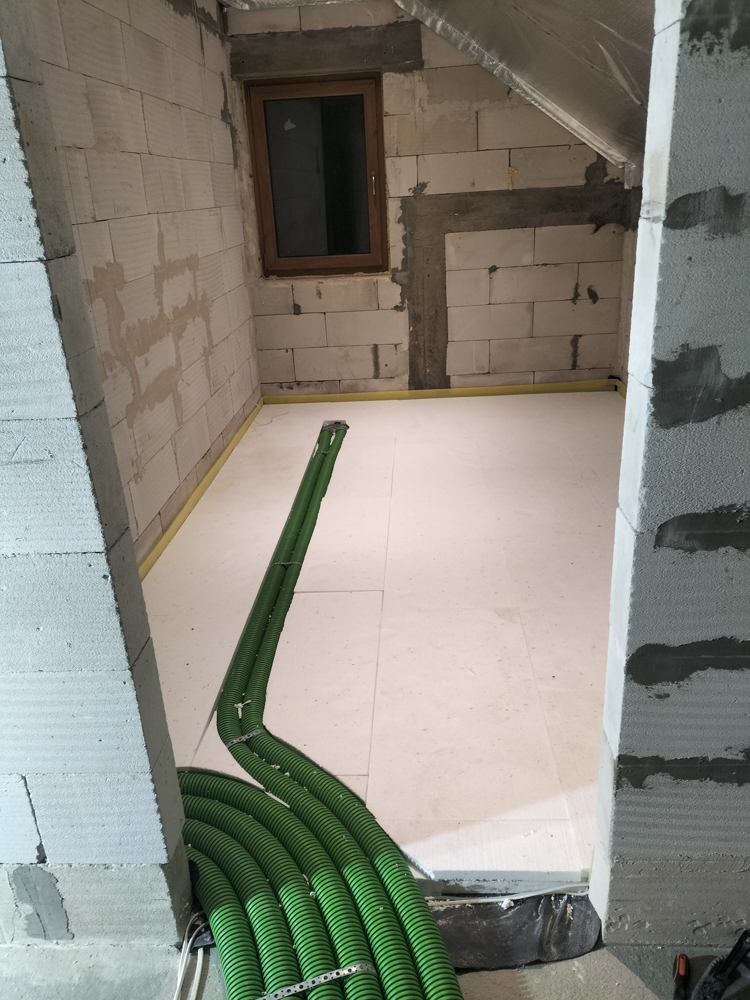 Izolacja podłogowa z zielonymi rurami ogrzewania podłogowego w surowym wnętrzu z bloczków. Widoczne okno i fragment poddasza.