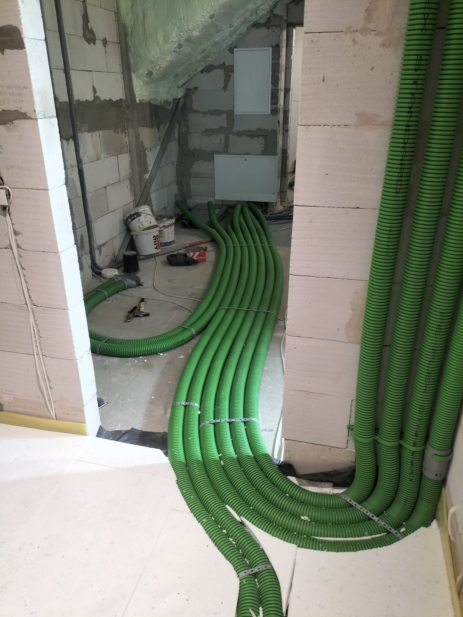 Zielone, karbowane rury instalacyjne ułożone wzdłuż ściany z białych bloczków, prowadzące do wnętrza budynku w trakcie budowy. Widoczne narzędzia i materiały budowlane.