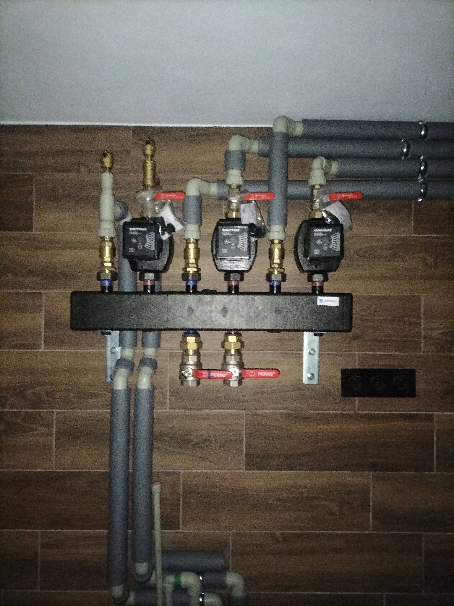 Rozdzielacz ogrzewania podłogowego z trzema pompami obiegowymi, zamontowany na ścianie z brązowych płytek imitujących drewno. Rury izolowane szarą pianką.
