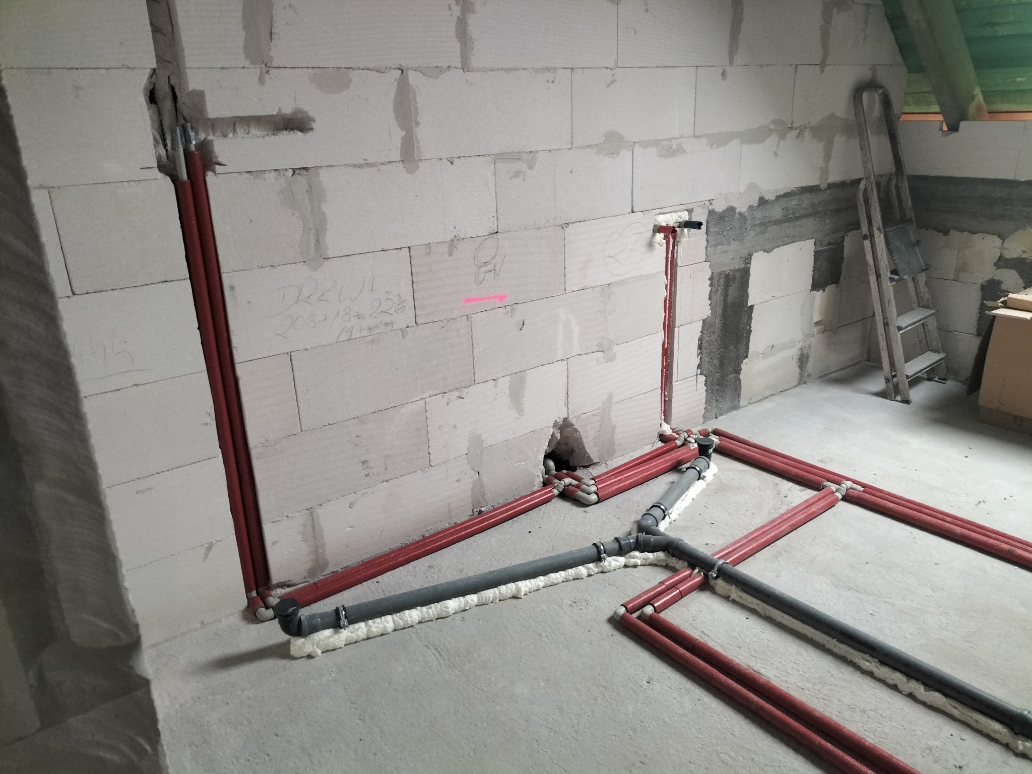Czerwone i szare rury instalacji wodnej/kanalizacyjnej ułożone na surowym betonie w pomieszczeniu z białymi ścianami z betonu komórkowego. Widoczna drabina.