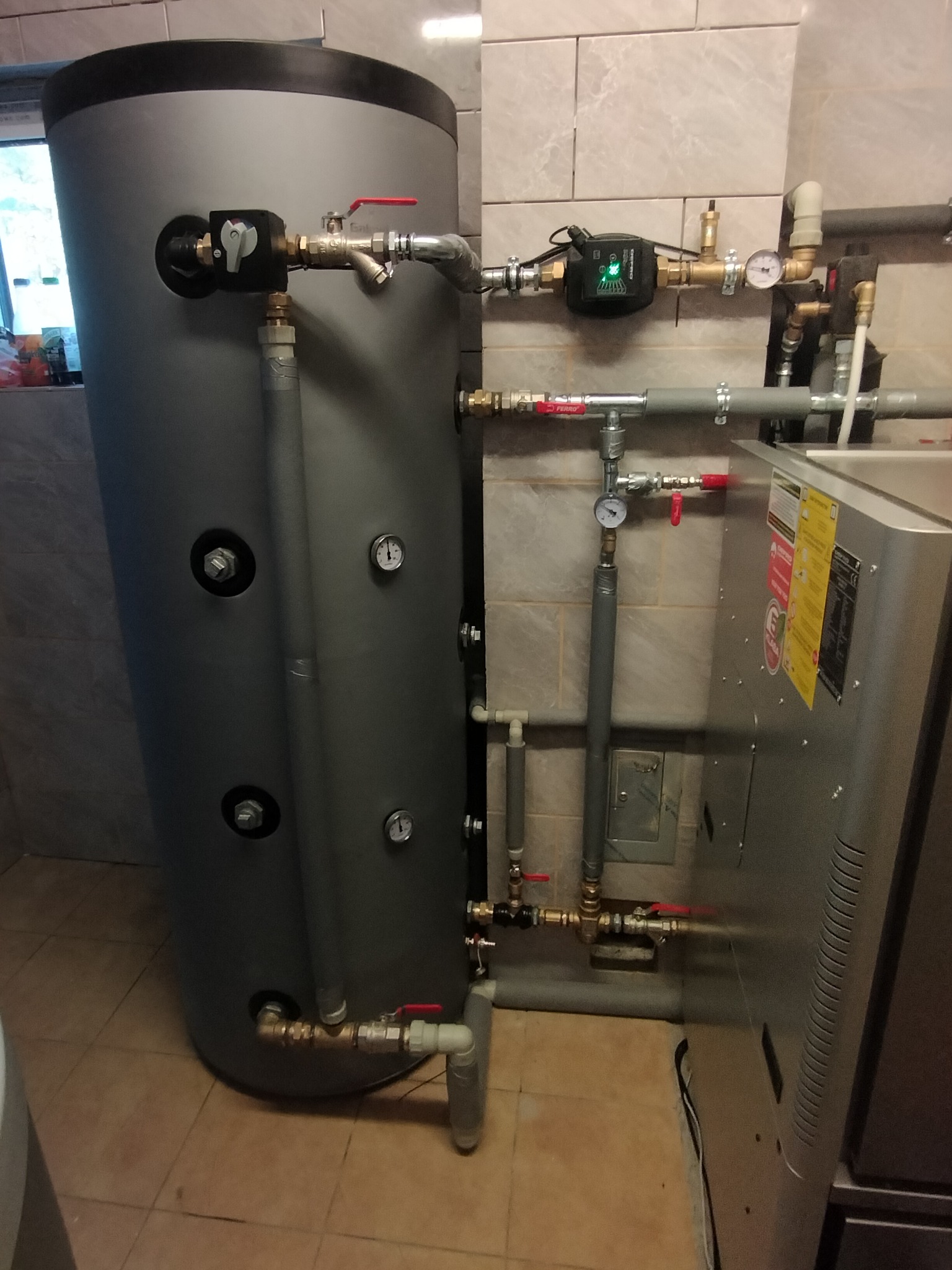 Szary, pionowy zbiornik buforowy z licznymi przyłączami rur, zaworami i manometrami w pomieszczeniu z beżowymi płytkami. Widoczny fragment pieca grzewczego.