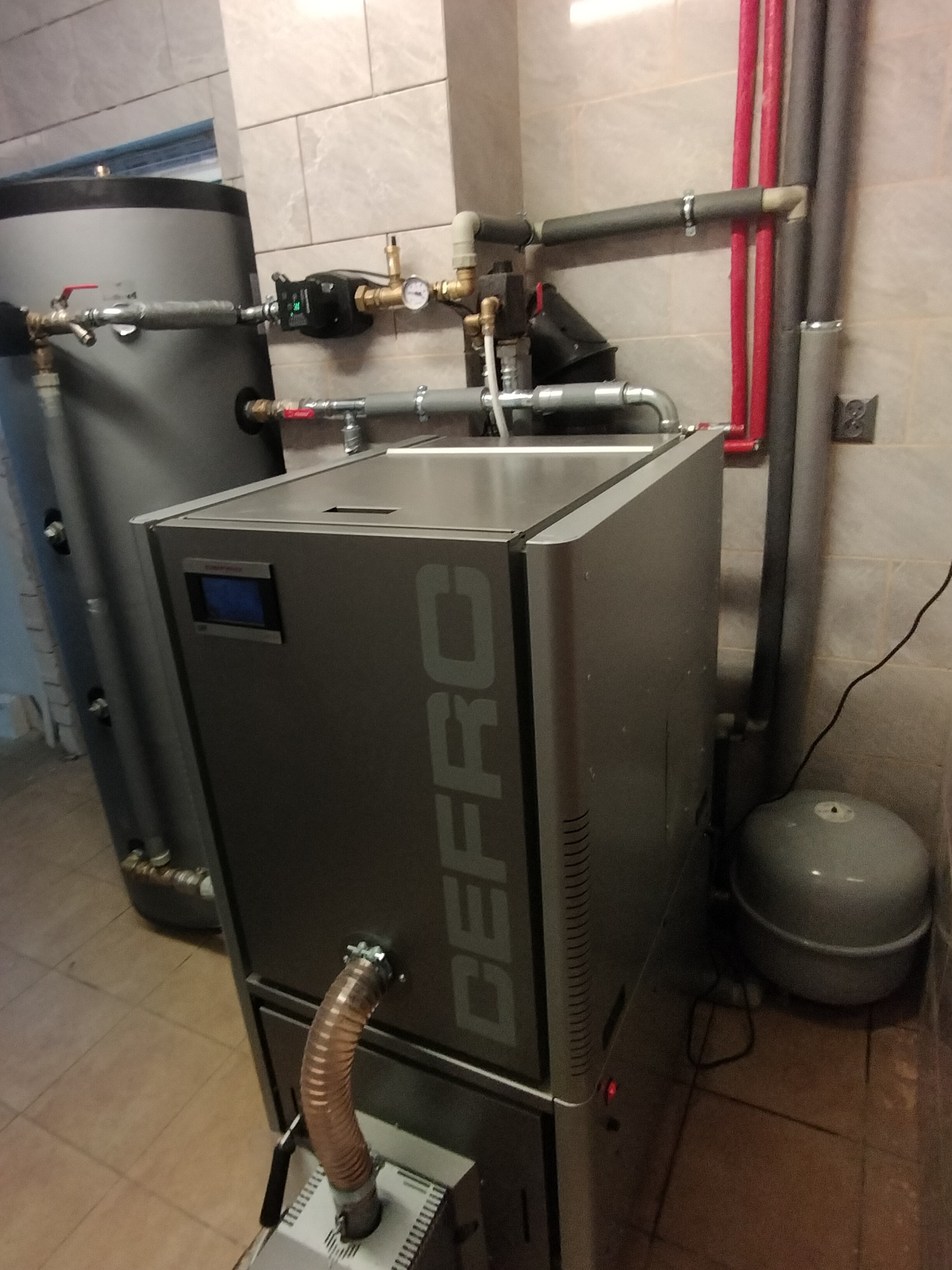 Nowoczesny piec CO marki DEFRO w kotłowni, z widocznymi rurami instalacji grzewczej i zasobnikiem ciepłej wody. Ujęcie z bliska, ukazujące detale podłączeń.