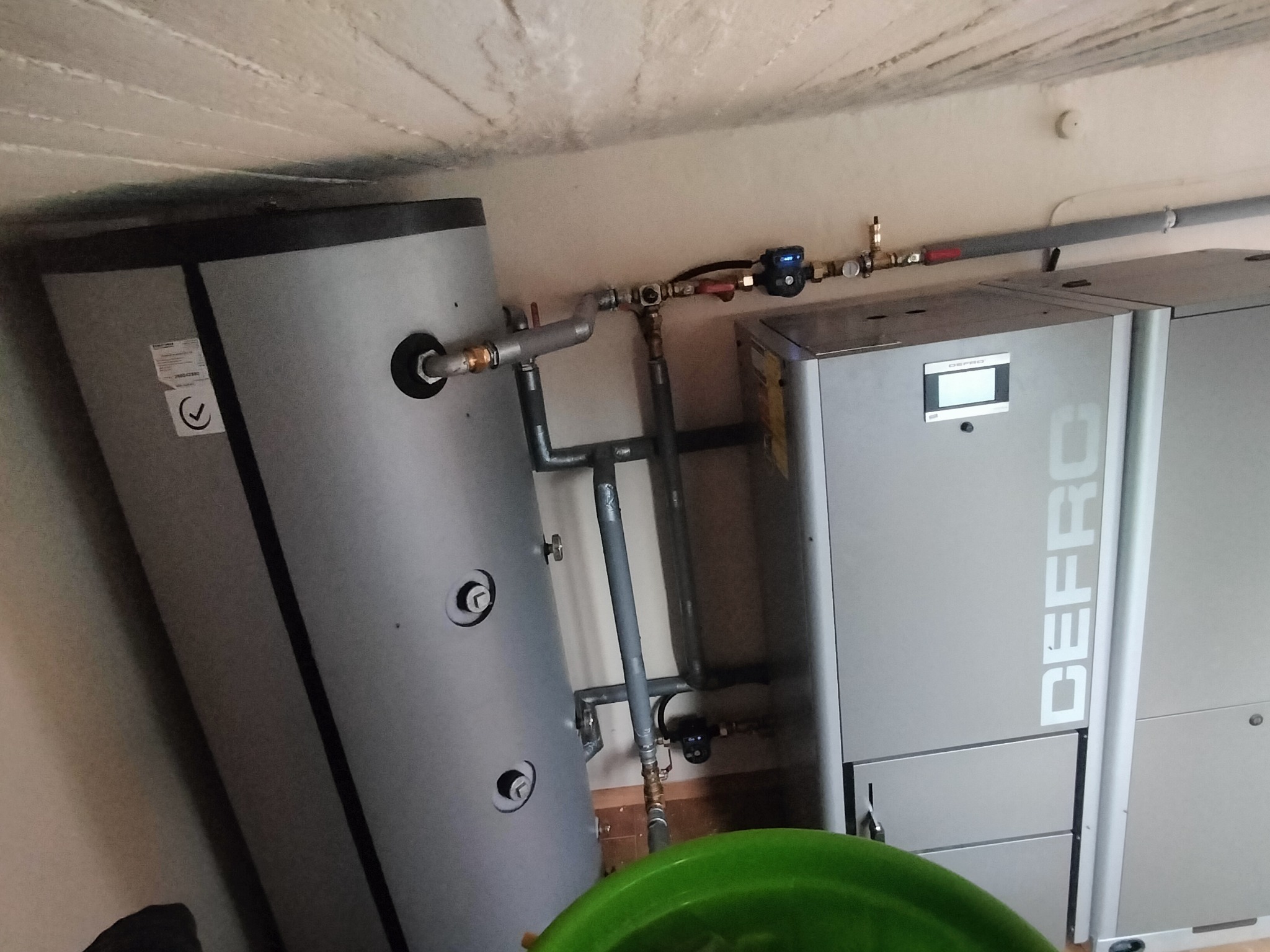 Szary piec CO marki Defro z podłączonym zasobnikiem wody i instalacją hydrauliczną. Widoczne rury, zawory i elementy sterujące. Ujęcie z dołu, fragment zielonego wiadra.