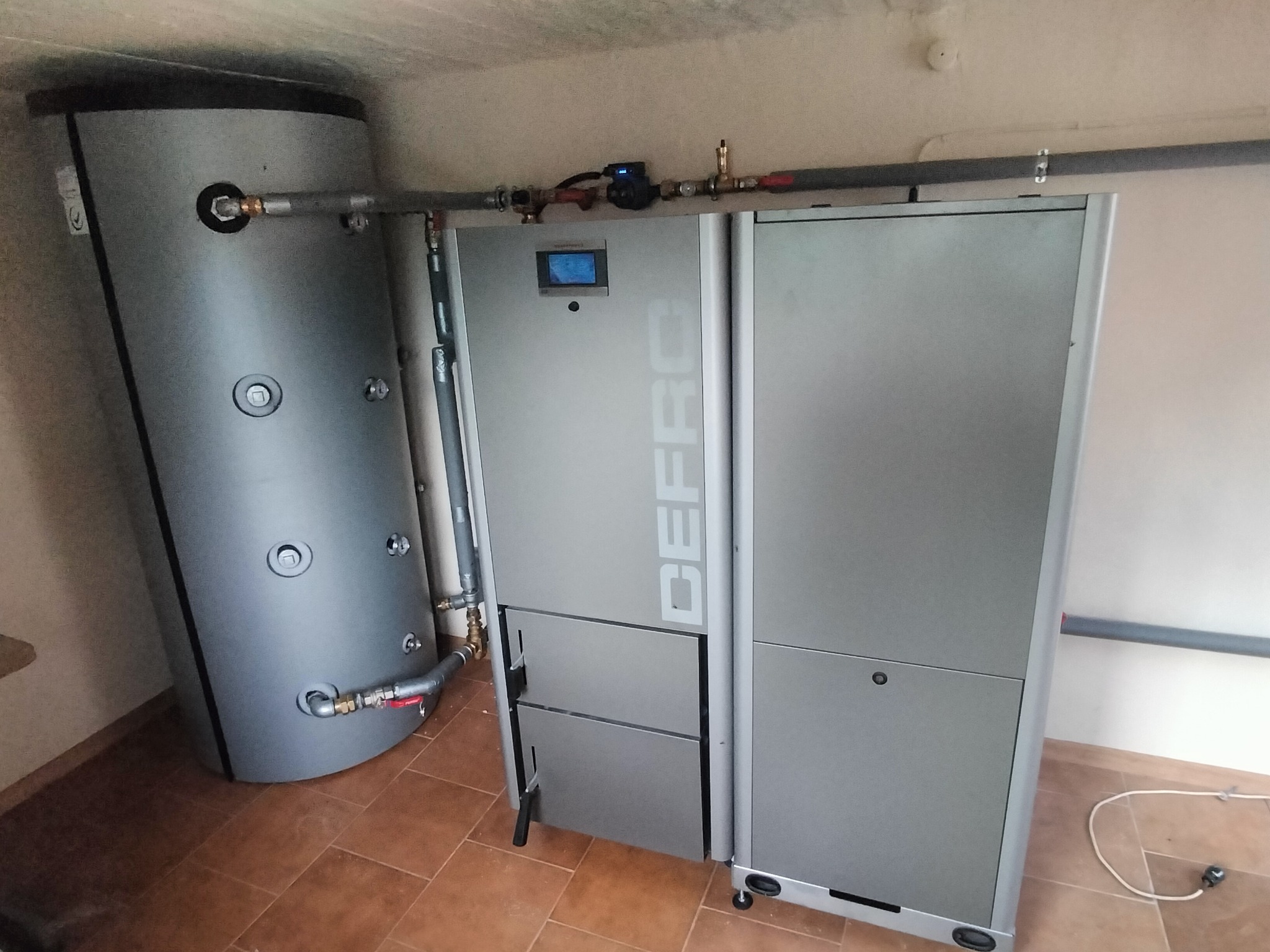 Szary piec CO marki DEFRO z podłączonym zasobnikiem wody, widoczne rury i armatura. Całość w pomieszczeniu z brązową terakotą.