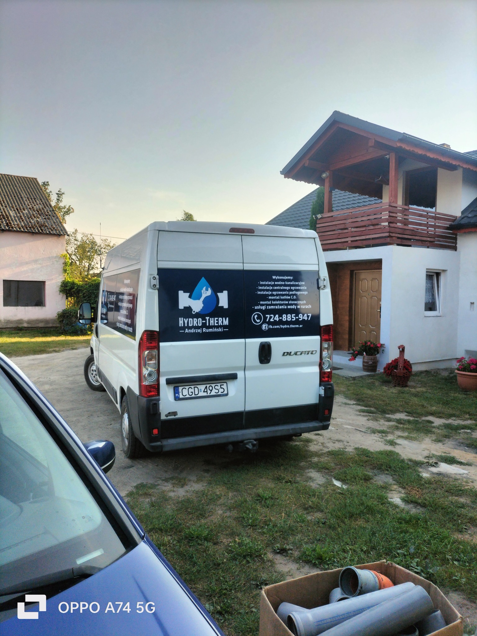 Biały bus firmy Hydro-Therm z logo i numerem telefonu na tle domu z drewnianym balkonem. W tle drugi budynek. W kadrze widoczne rury w kartonie.