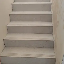 Wykończone schody wewnętrzne z szarymi, kamiennymi płytkami. Widoczne detale wykończenia stopni i ściany w trakcie prac. Kąt ujęcia z dołu schodów.