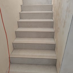 Usługi Ogólnobudowlane Piotr Mrotek - Wykończone schody z jasnych płytek z antypoślizgowymi paskami, widoczne ślady po pracach wykończeniowych na ścianach. Ujęcie z dołu schodów.