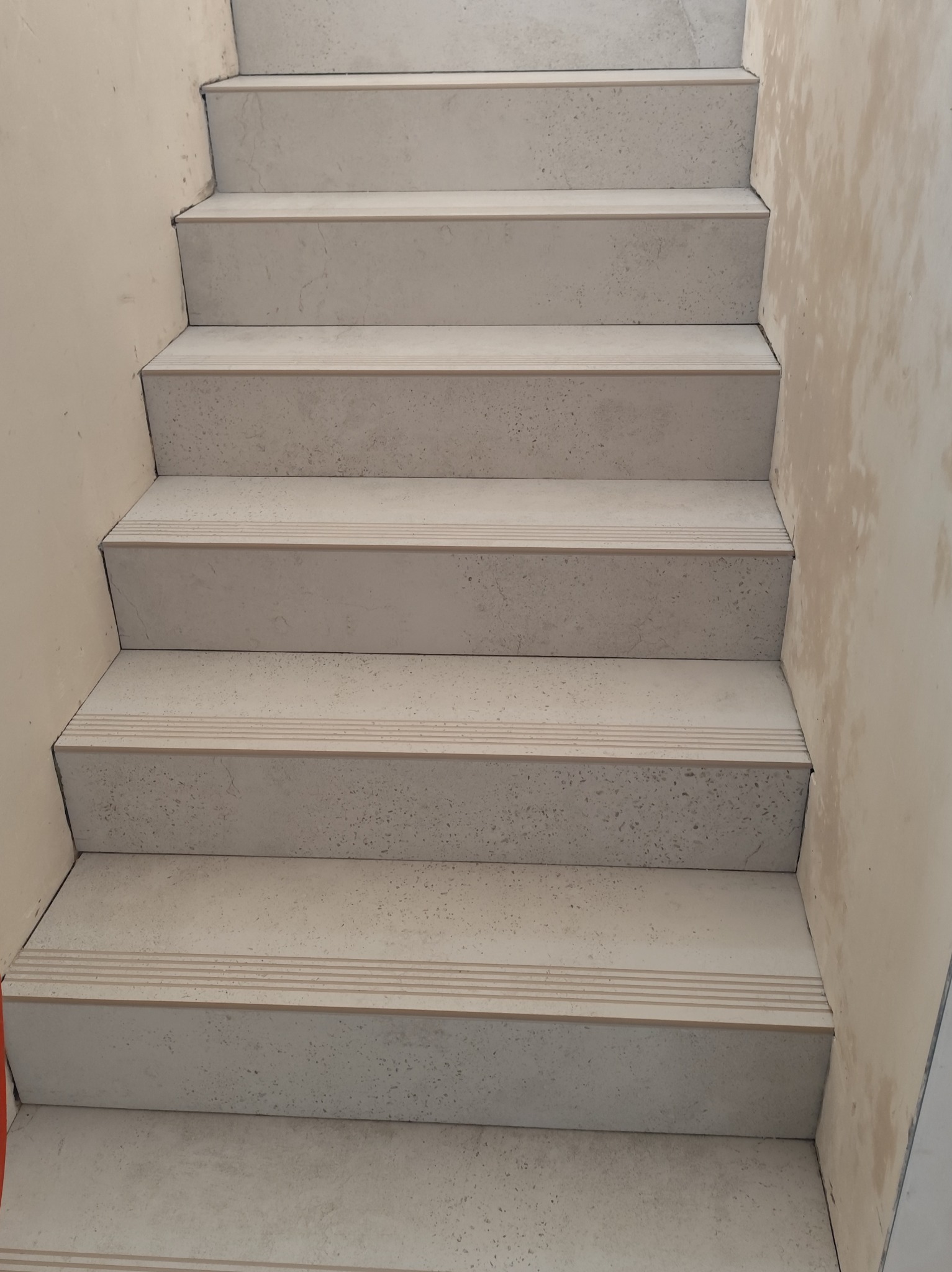 Wykończone schody wewnętrzne z szarymi, kamiennymi płytkami. Widoczne detale wykończenia stopni i ściany w trakcie prac. Kąt ujęcia z dołu schodów.