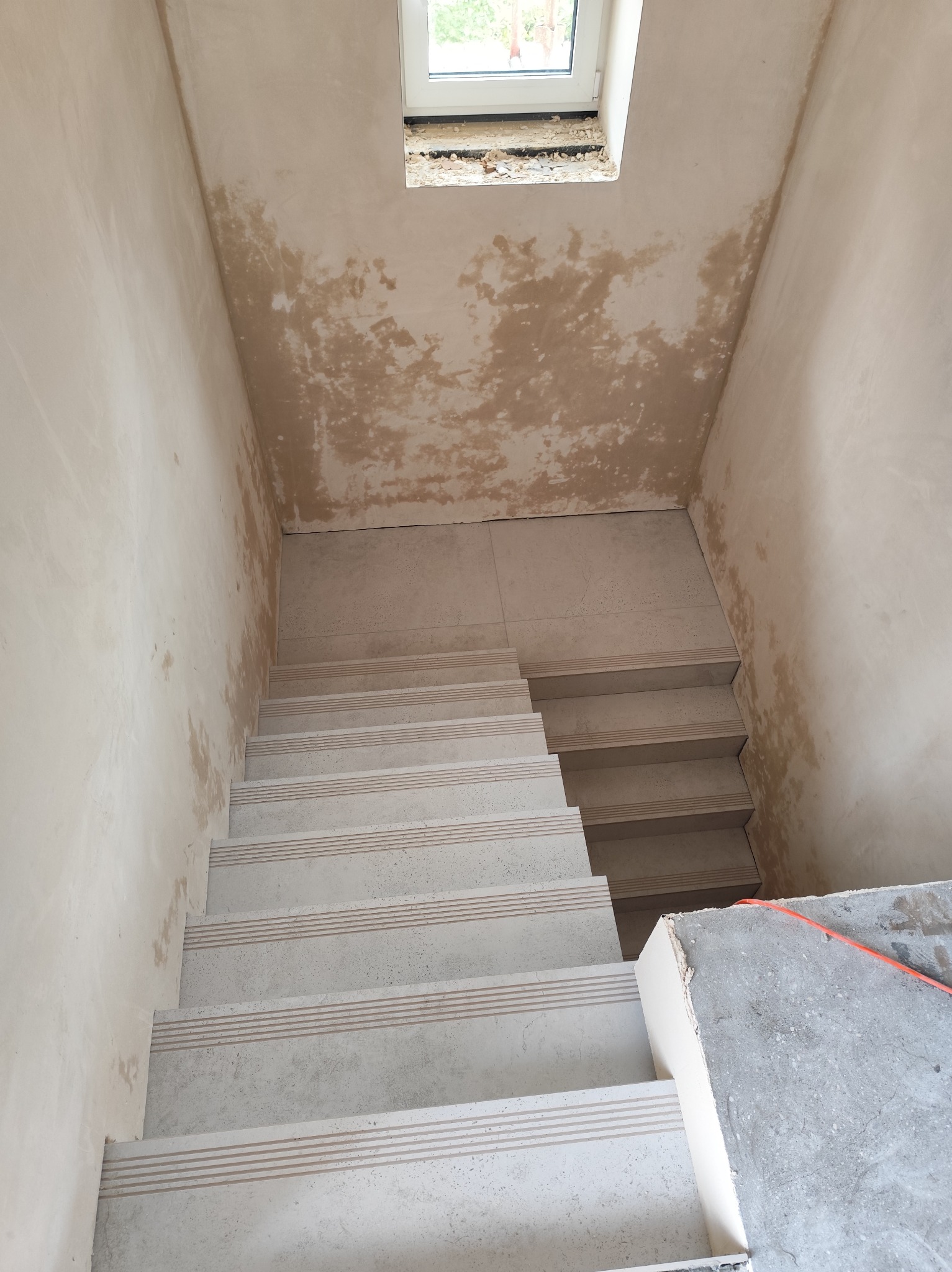 Wykończone schody z płytkami w jasnych odcieniach, widok z góry na tle ścian w trakcie szpachlowania. Małe okno w tle. Etap prac wykończeniowych.