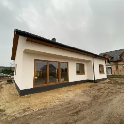 Niski, parterowy budynek z dużymi oknami w drewnianych ramach, wykończony na zewnątrz w kolorze białym, z czarnym dachem i podmurówką, otoczony ziemią i innymi budynkami w tle.