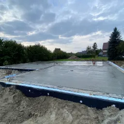 Świeżo wylana płyta fundamentowa domu jednorodzinnego z widoczną izolacją pionową, stalowymi prętami zbrojeniowymi wystającymi z betonu i otaczającym gruntem, w tle zabudowa jednorodzinna...
