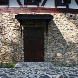 Wejście do budynku z kamienną elewacją i zadaszeniem nad drzwiami, widoczny fragment chodnika z kamiennej kostki