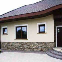 Elewacja domu jednorodzinnego z fragmentem ściany wykończonym kamieniem dekoracyjnym, widoczne trzy okna o brązowych ramach i wejście z trzema stopniami.