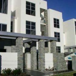 Nowoczesny budynek z elewacją w kolorze białym, z elementami wykończeniowymi z szarego kamienia, tworzącymi kolumny i fragmenty ścian. Widoczne duże, prostokątne okna i błękitne niebo w tle.