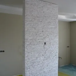 Ściana działowa wykończona białą mozaiką kamienną, widoczne otwory na gniazdka elektryczne i instalację hydrauliczną, podłoga zabezpieczona folią ochronną.