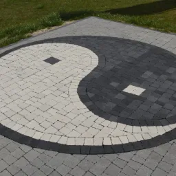 Nawierzchnia z kostki brukowej ułożona w kształt symbolu Yin i Yang, z wykorzystaniem jasnych i ciemnych odcieni szarości, otoczona szarą kostką i zielonym trawnikiem.
