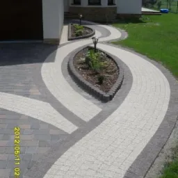 Kręty chodnik z jasnej kostki brukowej z ciemnoszarym obramowaniem i dwoma rabatami kwiatowymi z oświetleniem, prowadzący do domu z garażem.