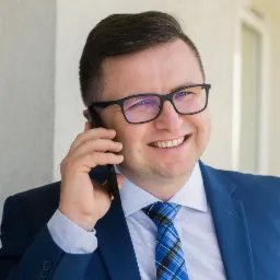 Uśmiechnięty mężczyzna w garniturze, z okularami, rozmawia przez telefon w Warszawie, na jasnym tle.