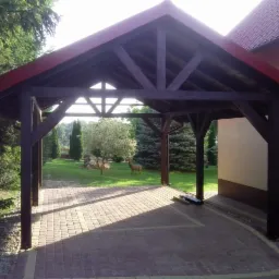 Drewniana wiata z czerwonym dachem na posesji z kostki brukowej, w tle trawnik z drzewami i dekoracyjnymi figurkami jeleni.