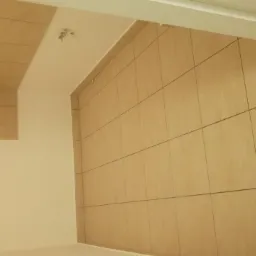 Beżowe płytki ceramiczne ułożone na ścianie i częściowo na suficie, widoczne fugi i oświetlenie sufitowe