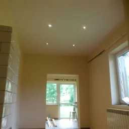 Wnętrze pokoju po remoncie z widocznym piecem kaflowym, świeżo pomalowanymi ścianami w odcieniach beżu, zamontowanymi punktowymi lampami sufitowymi i oknem z widokiem na zieleń.