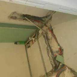 Fragment ściany i sufitu w trakcie prac: odsłonięte rury instalacji, częściowo zamontowana płyta karton-gips, butelka po napoju w tle.