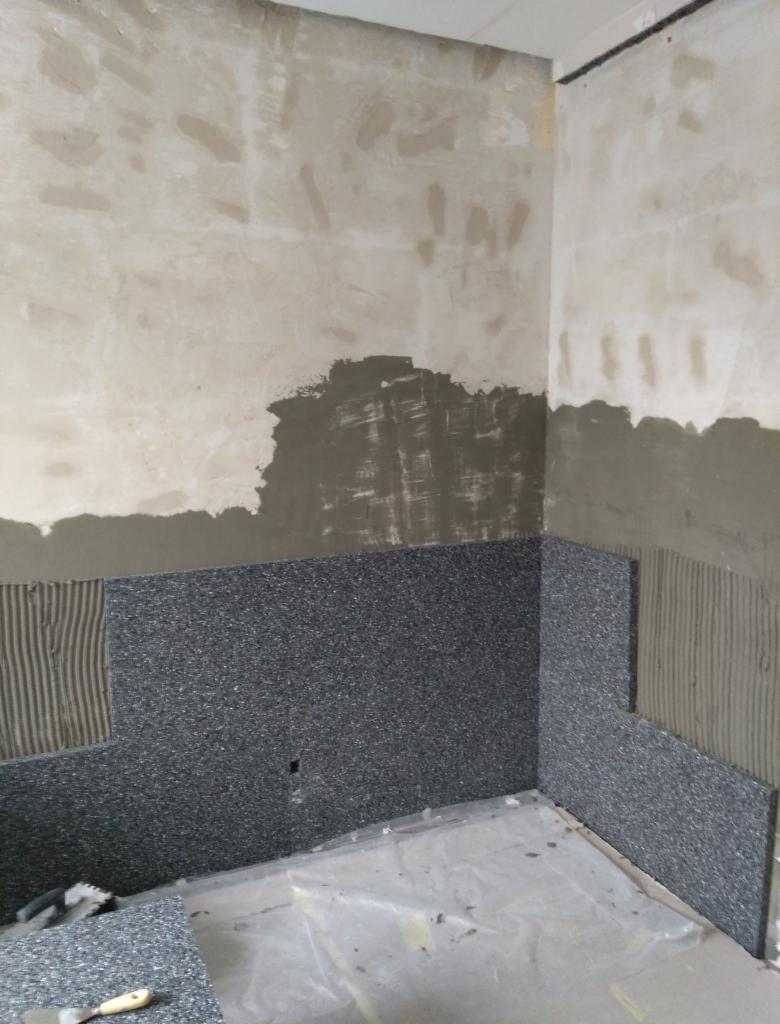 Proces układania ciemnych płytek na ścianie w pomieszczeniu, widoczny klej do płytek, szpachelka i folia ochronna na podłodze.