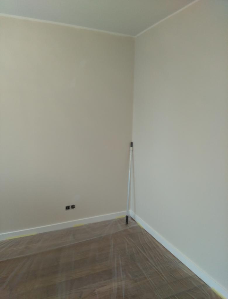 Świeżo pomalowane ściany w rogu pokoju, zabezpieczona podłoga folią, widoczny wałek malarski oparty o ścianę, gniazdka elektryczne na ścianie.