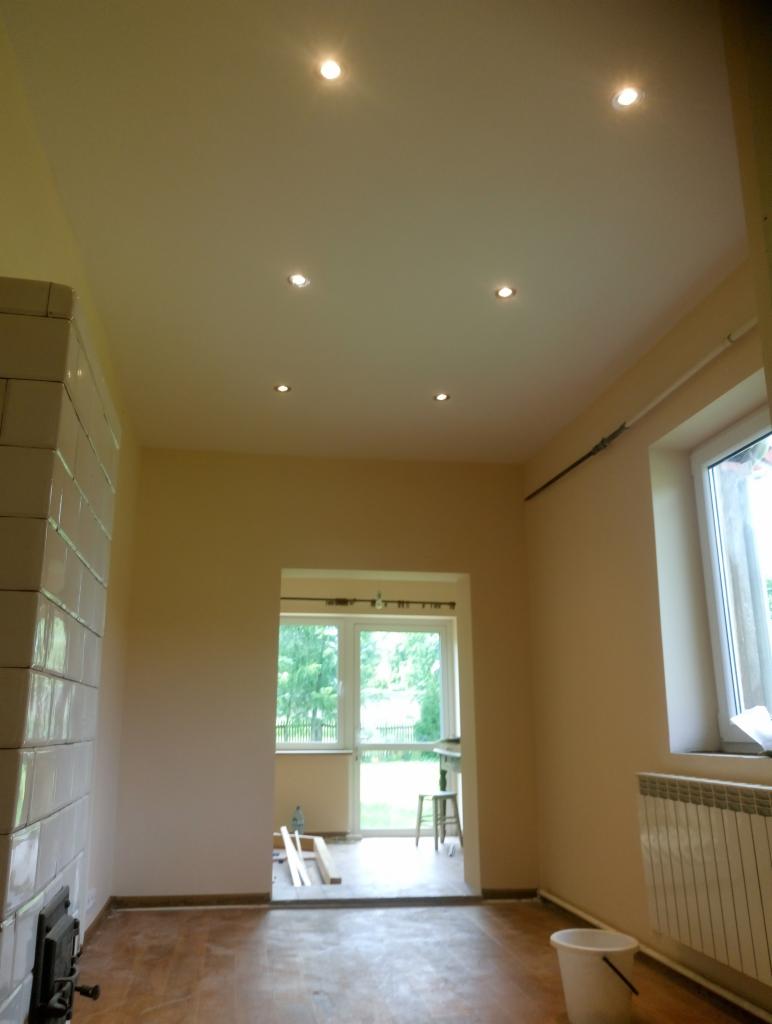 Wnętrze pokoju po remoncie z widocznym piecem kaflowym, świeżo pomalowanymi ścianami w odcieniach beżu, zamontowanymi punktowymi lampami sufitowymi i oknem z widokiem na zieleń.