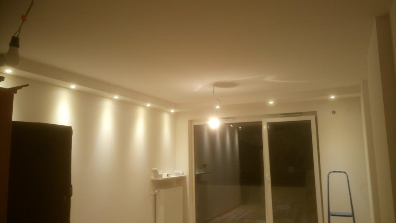 Wykończone wnętrze pokoju z oświetleniem punktowym w suficie podwieszanym i wiszącą żarówką na kablu, widoczne przesuwne drzwi balkonowe i drabina malarska.