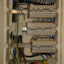 Otwarta skrzynka elektryczna z bezpiecznikami, przewodami i listwami zaciskowymi. Widoczne różne kolory przewodów i elementy montażowe.