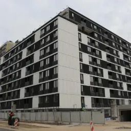 Wielopiętrowy budynek mieszkalny w trakcie budowy, z widocznymi rusztowaniami i pracownikami w kamizelkach odblaskowych, teren budowy zabezpieczony metalowym ogrodzeniem.