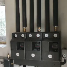 Rozdzielacz ogrzewania podłogowego z trzema obiegami, każdy z pompą obiegową i manometrem, oraz izolowane rury doprowadzające, zamontowany na ścianie w surowym stanie.