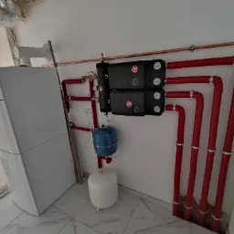 Kotłownia w trakcie montażu: piec kondensacyjny w folii ochronnej, czerwone rury grzewcze, zasobnik ciepłej wody i naczynia przeponowe, instalacja w surowym stanie.