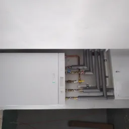 Biała pompa ciepła Viessmann zamontowana na ścianie, z widocznymi rurami miedzianymi i izolowanymi szarymi rurami instalacji grzewczej oraz zaworami. Widoczne elementy surowego wnętrza w tle.