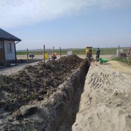 MATEUSZ BEZWERCHNY INSTALACJE - Wykop pod rury instalacyjne w ziemi, widoczny budynek mieszkalny w tle, na budowie obecny sprzęt budowlany i pracownicy.