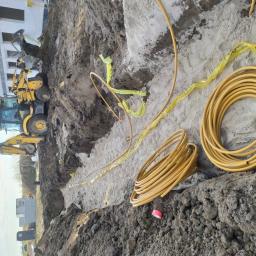 MATEUSZ BEZWERCHNY INSTALACJE - Wykop z ułożonymi żółtymi rurami i taśmą ostrzegawczą, na budowie w tle widoczna koparka JCB.