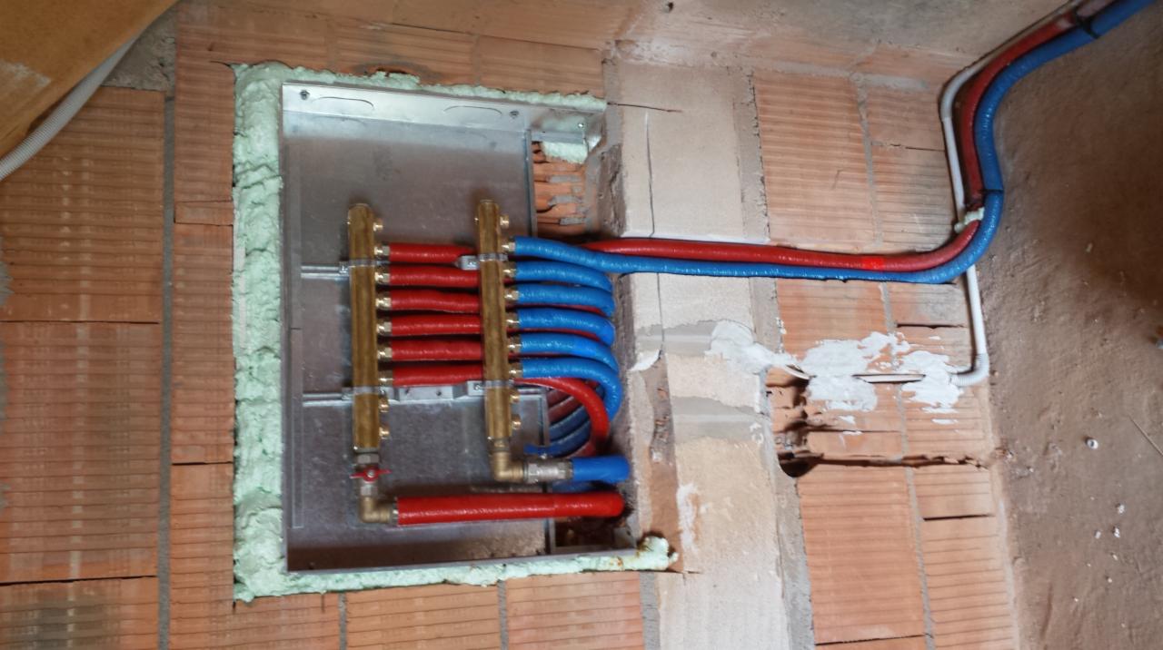 Rozdzielacz instalacji grzewczej z czerwonymi i niebieskimi rurami w ścianie z cegły, uszczelniony pianką montażową.