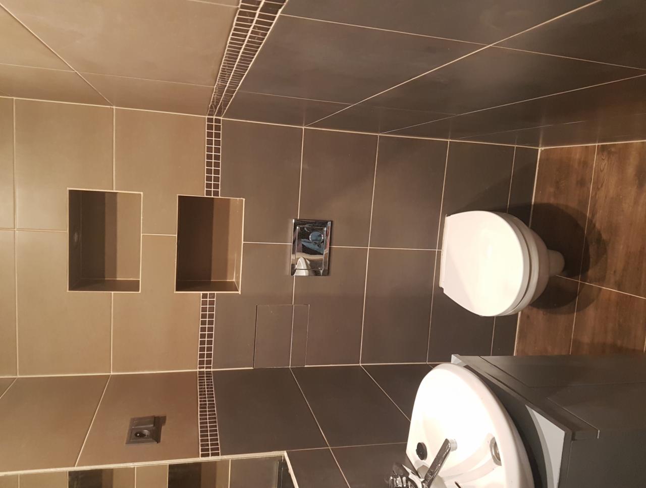 Wykończona łazienka w odcieniach brązu i beżu z dekoracyjnymi wstawkami mozaiki. Widoczna toaleta, umywalka z szafką i wnęki w ścianie.