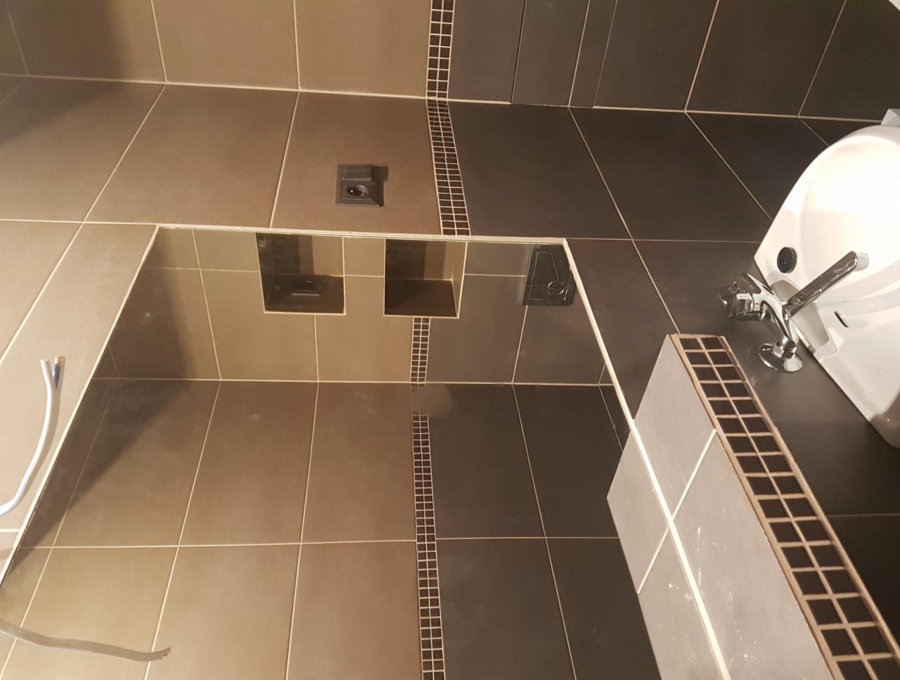 Wykończona łazienka z brązowo-szarymi płytkami, widoczny odpływ liniowy, umywalka z baterią i fragment wężyka doprowadzającego wodę.