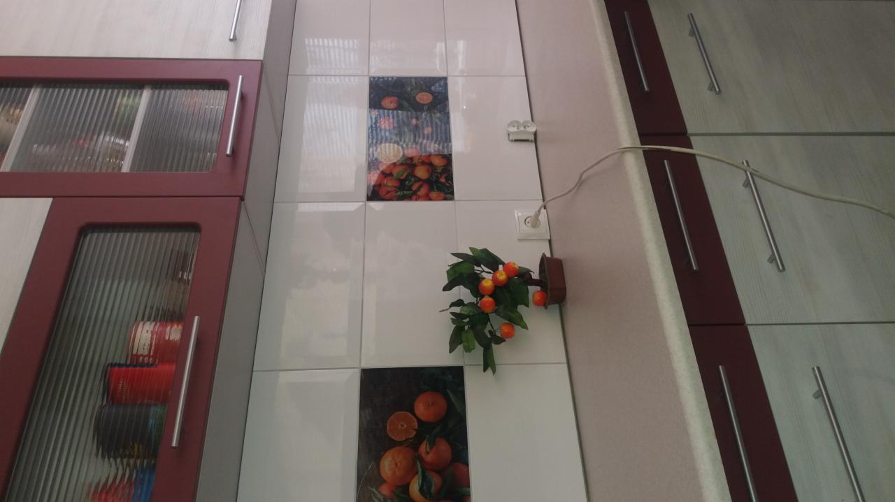 Białe płytki ścienne z motywem owoców cytrusowych, ozdobna roślina z pomarańczowymi owocami, fragment szafek kuchennych z ryflowanymi szybami i podłużnymi uchwytami.