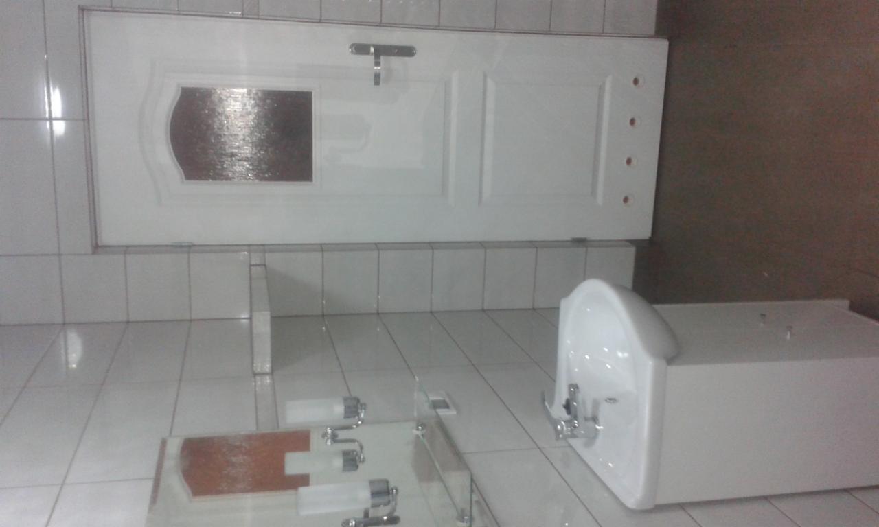 Biała umywalka z baterią, zamontowana w łazience wyłożonej białymi płytkami, obok drzwi z szybką i klamką