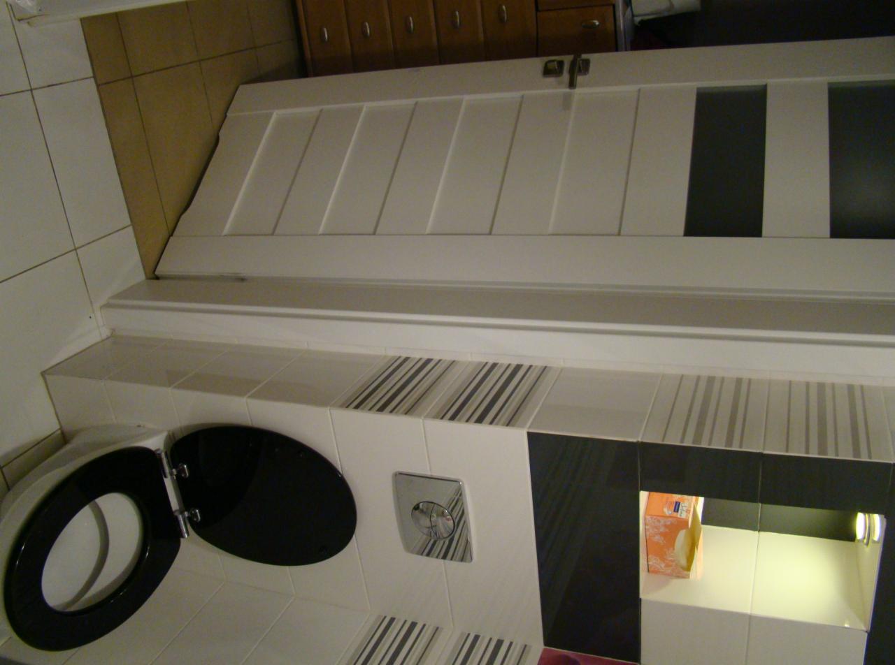 Wykończona łazienka w Warszawie z białą zabudową geberit, czarną deską sedesową i akcentami w postaci płytek w paski oraz podświetlanej wnęki.