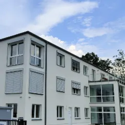 Biały budynek biurowy z szarymi oknami i roletami zewnętrznymi, widok z dołu na tle błękitnego nieba z chmurami.