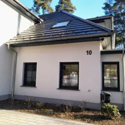 Nowoczesny dom jednorodzinny z czarnymi oknami i numerem domu 10, widok z zewnątrz na elewację z elementami dachu i ogrodu.