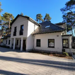 Elewacja nowoczesnego, dwukondygnacyjnego domu z białym tynkiem, czarną stolarką okienną i dachem z ciemnej dachówki, numer 10 na ścianie, otoczonego wysokimi sosnami i brukowanym podjazdem...