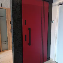 Czerwone drzwi wejściowe z czarnymi, pionowymi paskami i długim, prostokątnym uchwytem, osadzone w czarnej ościeżnicy z dekoracyjną tapetą w tle, widoczne obok innych drzwi.