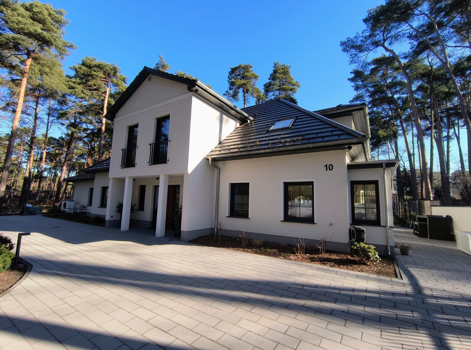 Elewacja nowoczesnego, dwukondygnacyjnego domu z białym tynkiem, czarną stolarką okienną i dachem z ciemnej dachówki, numer 10 na ścianie, otoczonego wysokimi sosnami i brukowanym podjazdem...