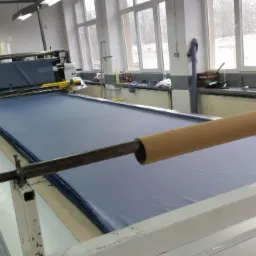 Warstwy niebieskiego materiału rozłożone na stole krojczym z automatycznym nożem, widok z boku na jasne wnętrze pracowni.